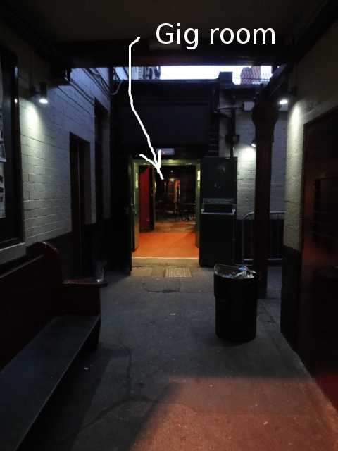 Photo: a small courtyard with concrete floor, with a wooden bench on the left. Ahead about 4 or 5 metres is an open door leading to inside the building.
